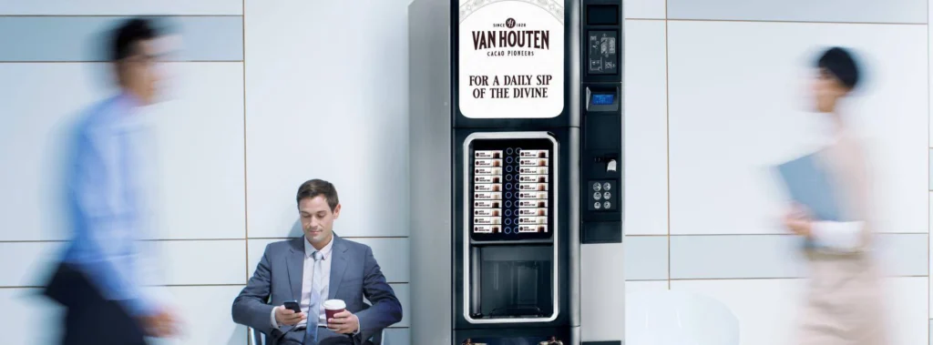 Elegancki mężczyzna pije gorącą czekoladę obok nowoczesnego automatu vendingowego Van Houten w profesjonalnym otoczeniu biurowym.