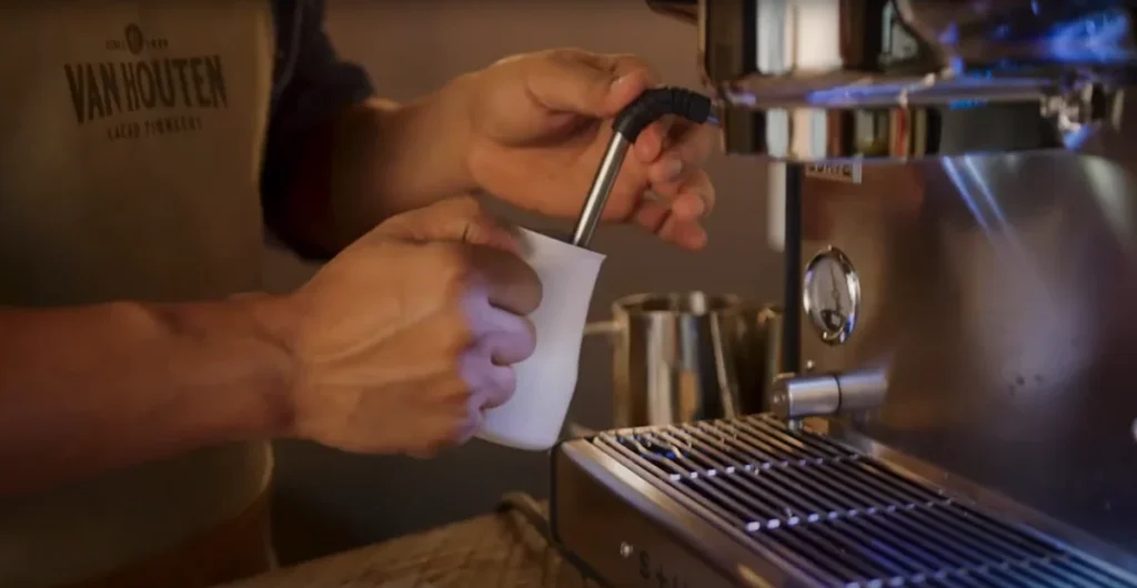 Barista w koszulce z logo Van Houten przygotowuje gorącą czekoladę na profesjonalnym ekspresie ciśnieniowym w kawiarni.